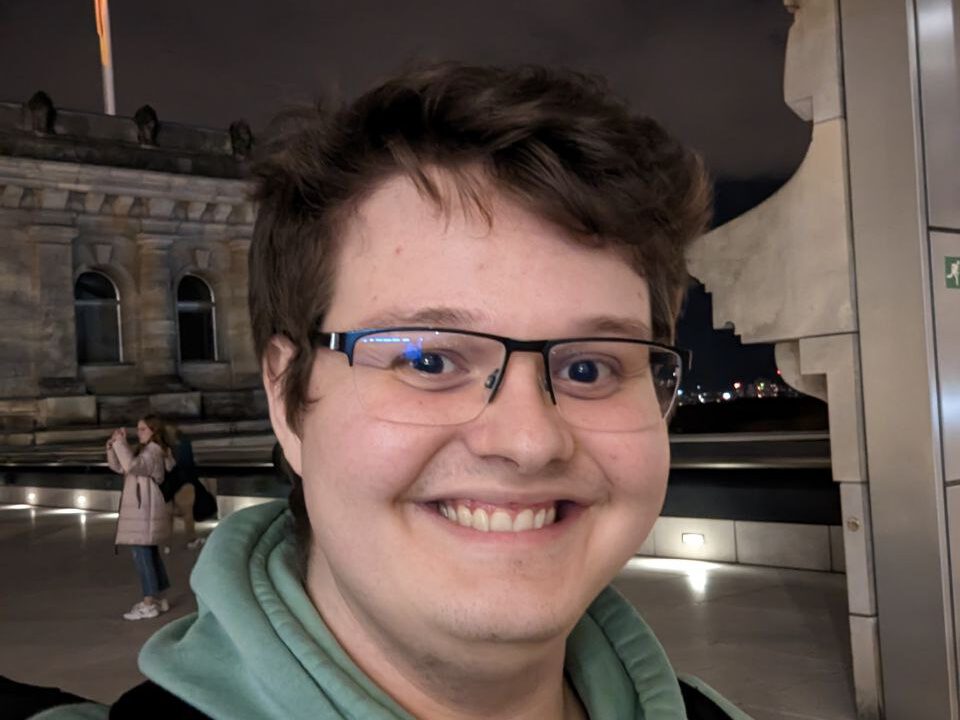 Dieses Bild ist ein abends aufgenommenes Selfie und zeigt Lorenzo lachend bei der Reichstagskuppel. Er hat kurze braune Haare, trägt eine Brille und einen hellgrünen Kapuzenpullover.